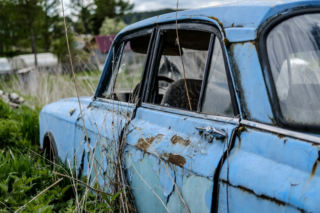 a heavily rusted and deteriorating blue car abandoned in tall grass